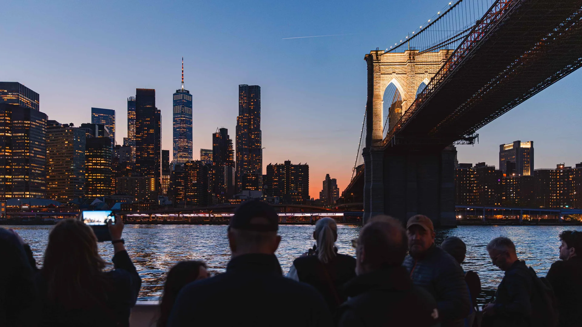 Guests on a Circle Line deck at dusk under the Brooklyn Bridge with the downtown skyline, from the harbor lights cruise listing